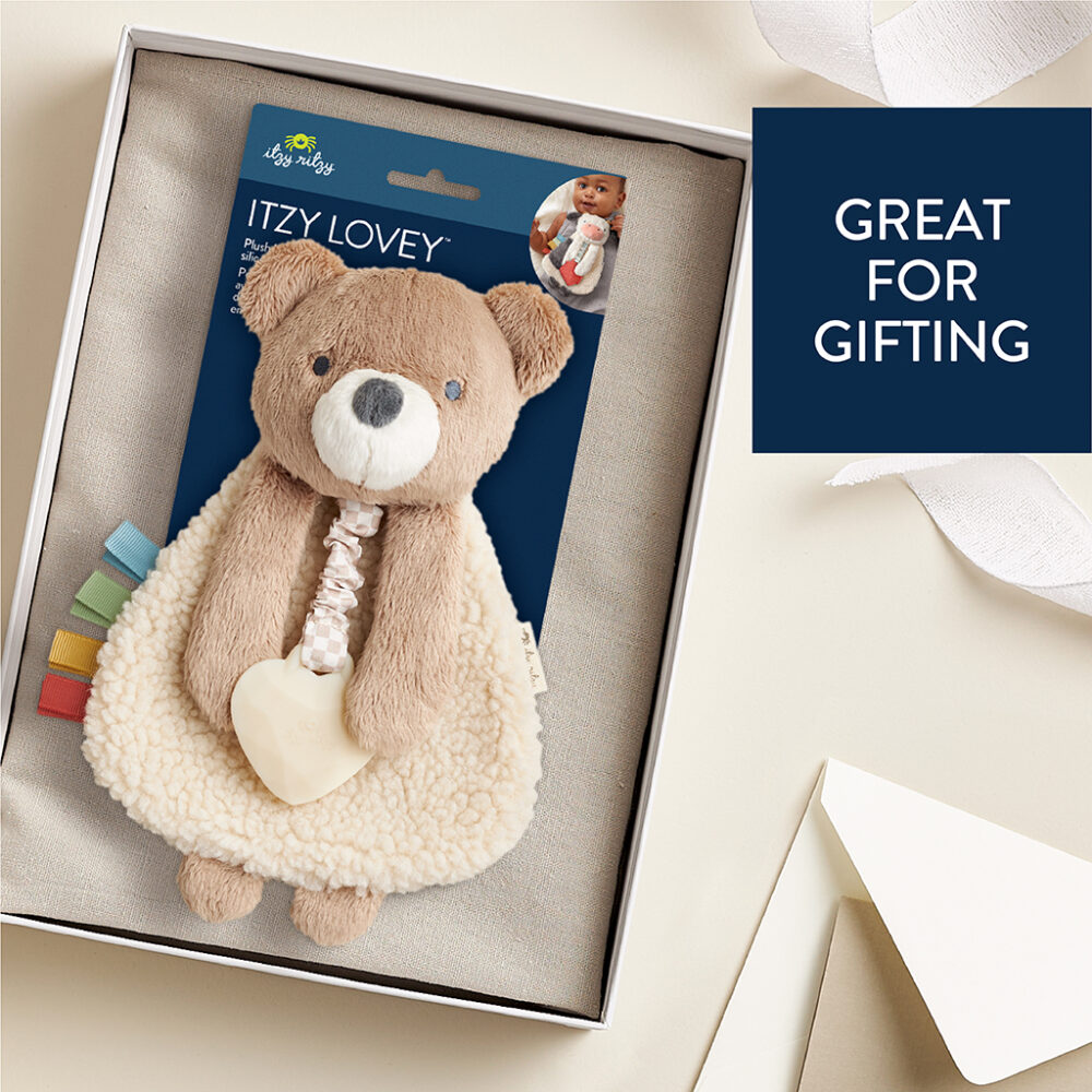 A plush teddy bear blanket with colorful tabs, packaged in a box. A tag reads "Itzy Lovey." A label on the side reads "Great for Gifting.