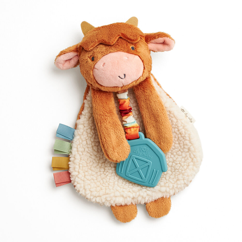 A plush cow toy with a soft, brown and cream body, holding a blue textured teether shaped like a barn. The toy features colorful fabric tags on one side.