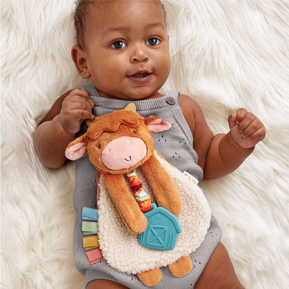 A baby in a gray onesie lying on a white fluffy surface holds a plush cow toy with various textures and colors.