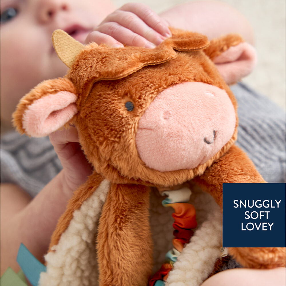 A baby holds a soft, plush cow toy labeled "SNUGGLY SOFT LOVEY.