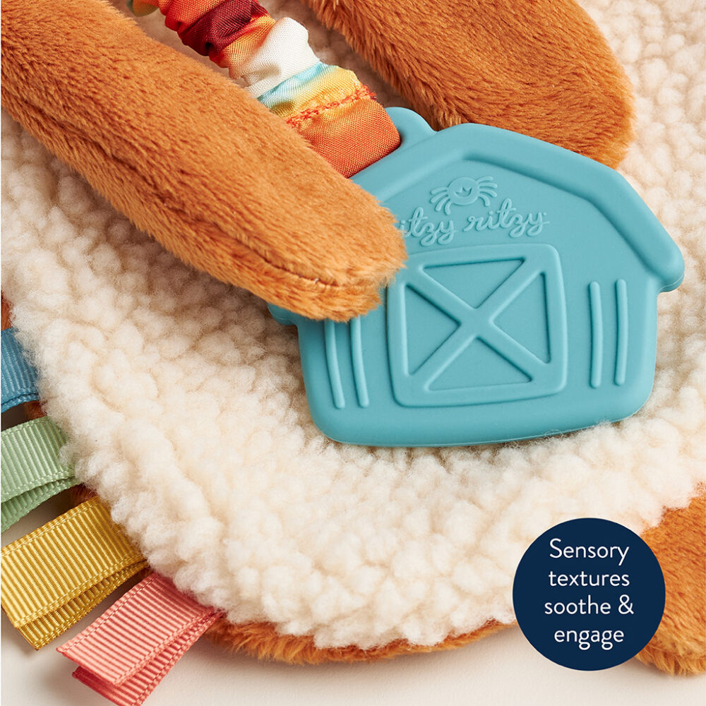 Close-up of a sensory toy with various textures, including a soft white fabric, colorful ribbons, and a blue rubber barn-shaped piece with "Itzy Ritzy" text. Round label reads: "Sensory textures soothe & engage.