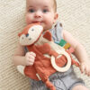 A baby lying on a cream-colored textured blanket, holding a plush fox toy with colorful tabs and a teething ring. The baby is wearing a light gray knitted outfit.