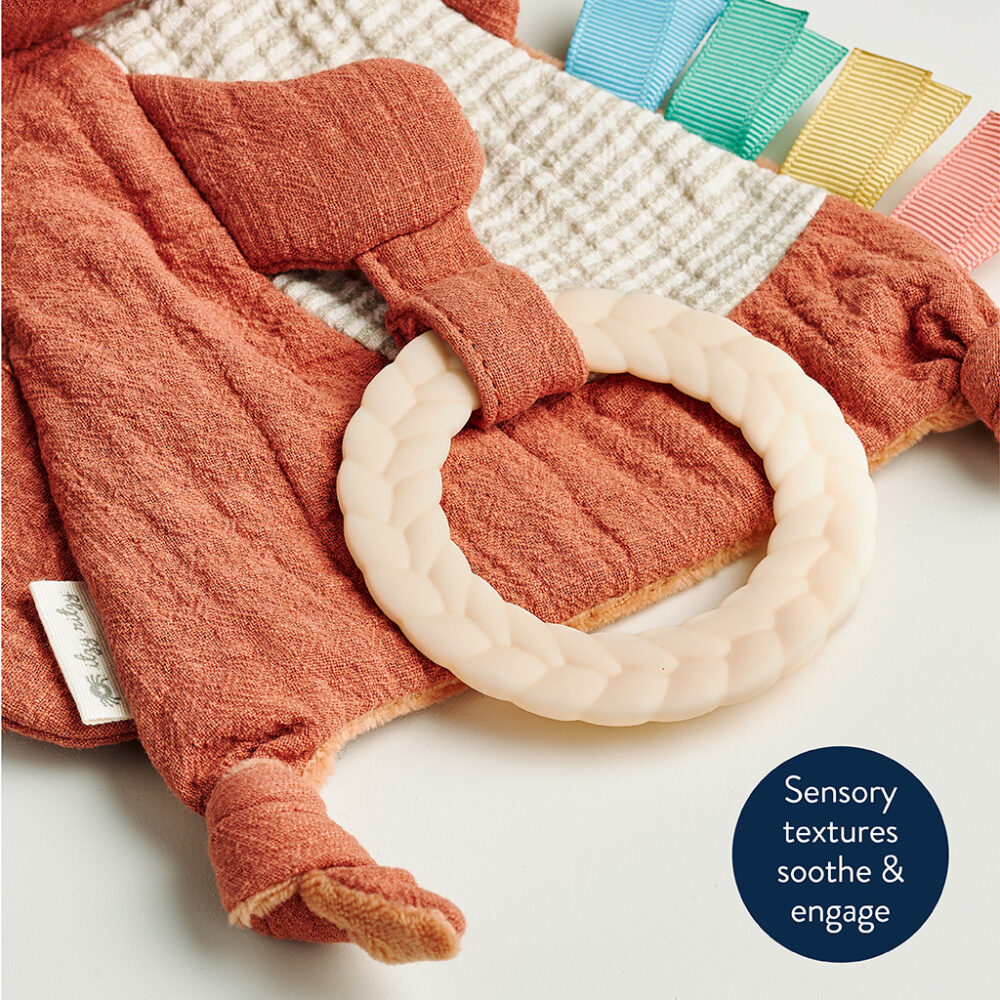 A sensory baby toy with a braided teething ring and various fabric textures, including a white and orange checkered cloth and colorful ribbon tags. Text reads: "Sensory textures soothe & engage.