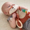 A baby lies on a textured beige surface, holding and chewing on a soft, stuffed toy designed to resemble an animal with colorful fabric tabs.