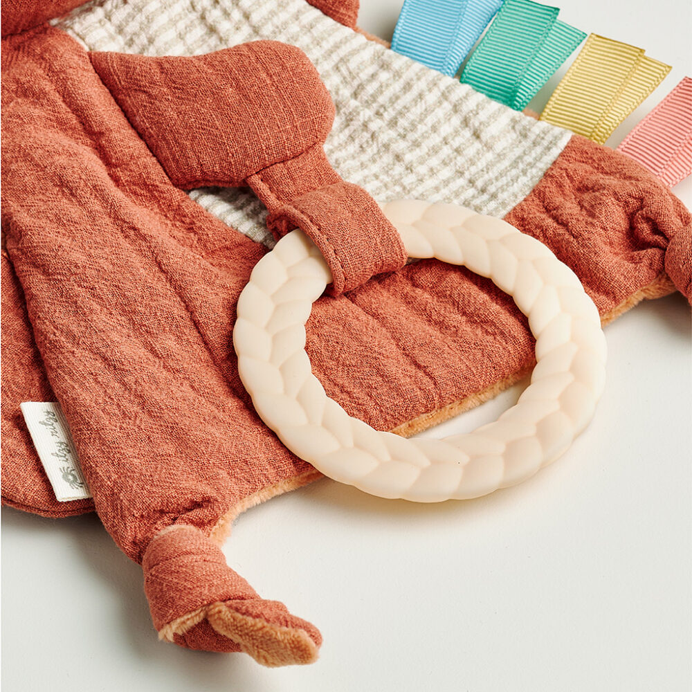 A soft fabric baby toy with a textured teething ring and multiple colorful tags attached. The toy features a mix of terracotta and white waffle fabric.