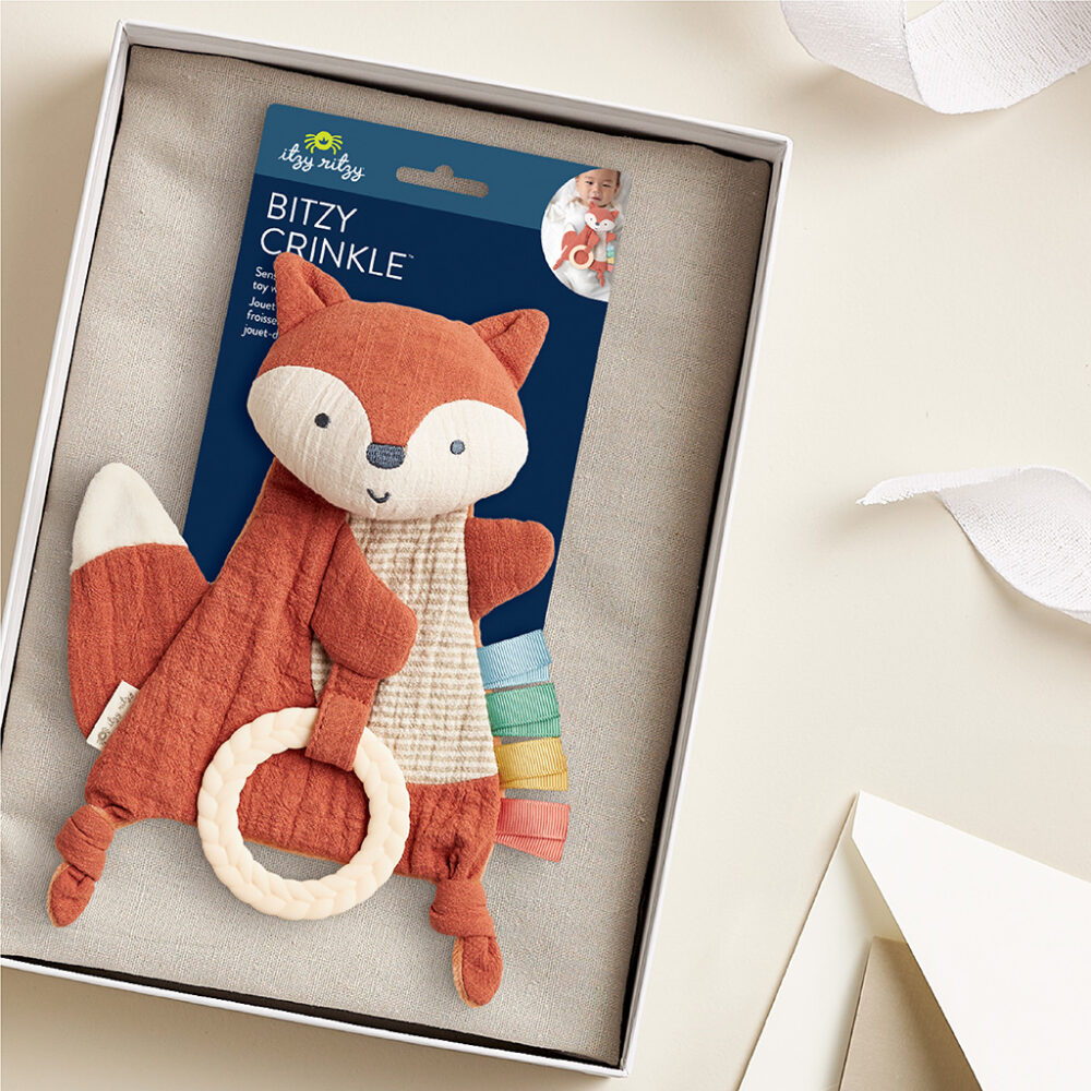 A boxed baby toy featuring a fox with a textured ring, labeled "Bitzy Crinkle" by Itzy Ritzy, surrounded by a white ribbon and paper.