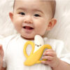 A baby in a white outfit holds an Itzy Ritzy Chew Crew Silicone Teether with Handles, shaped like a yellow and white banana with a smiling face, while sitting on a fluffy white surface.