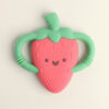 The Itzy Ritzy Chew Crew Silicone Teether with Handles is a strawberry-shaped baby teether featuring a smiling face and two green handles designed for easy gripping.