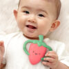 Infant lying on a white surface, holding an Itzy Ritzy Chew Crew Silicone Teether with Handles shaped like a strawberry, and smiling.