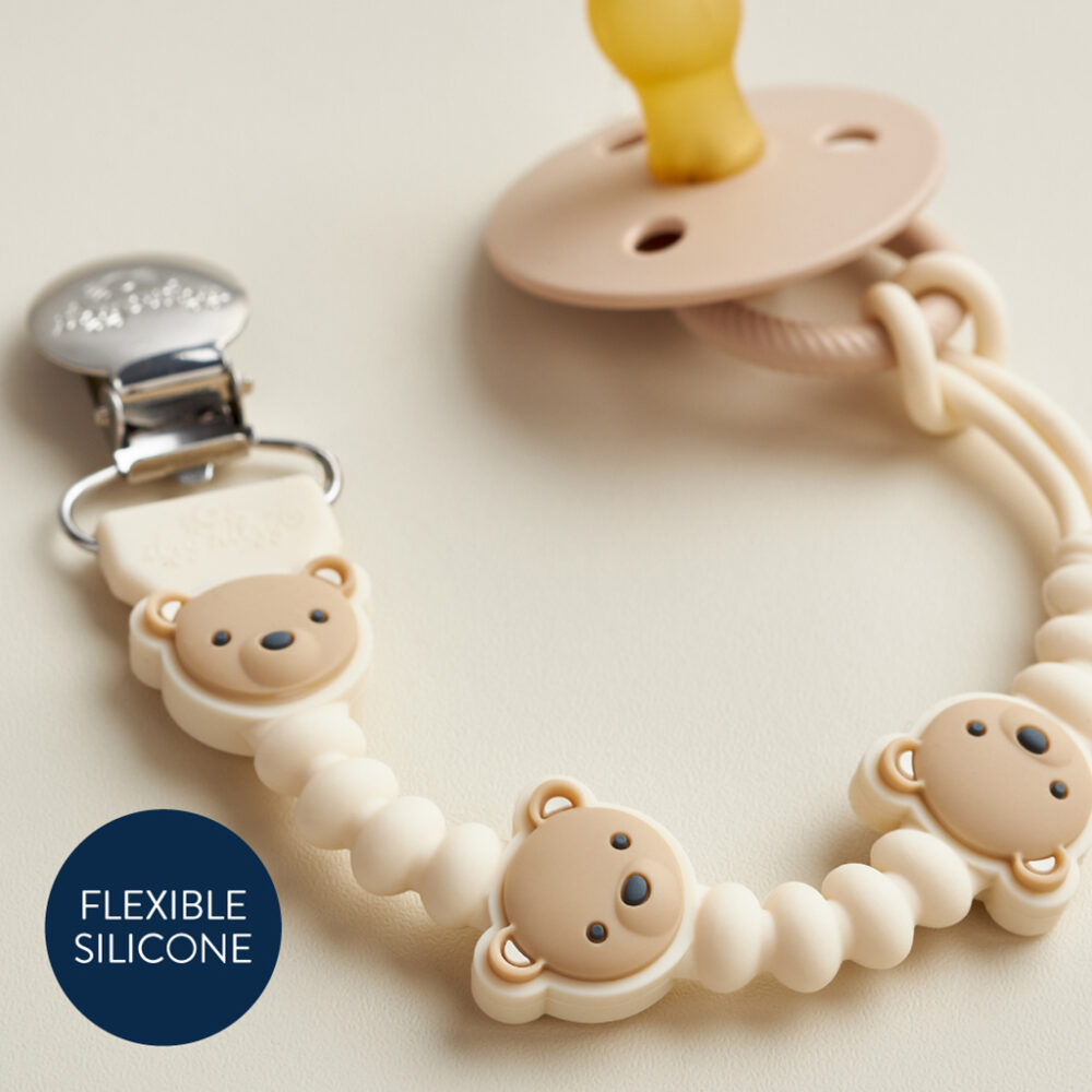 The Itzy Ritzy Sweetie Strap Plus Bear Silicone Pacifier Clip in beige, featuring a bear-themed silicone bead clip, is lying on a light surface with a label that reads "Flexible Silicone.