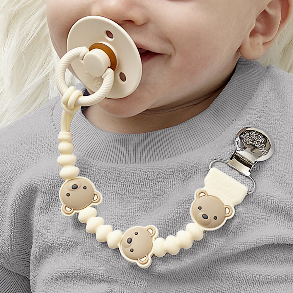 A baby with an Itzy Ritzy Sweetie Strap Plus Bear Silicone Pacifier Clip attached to their gray shirt, featuring bear-shaped beads linking to the pacifier.