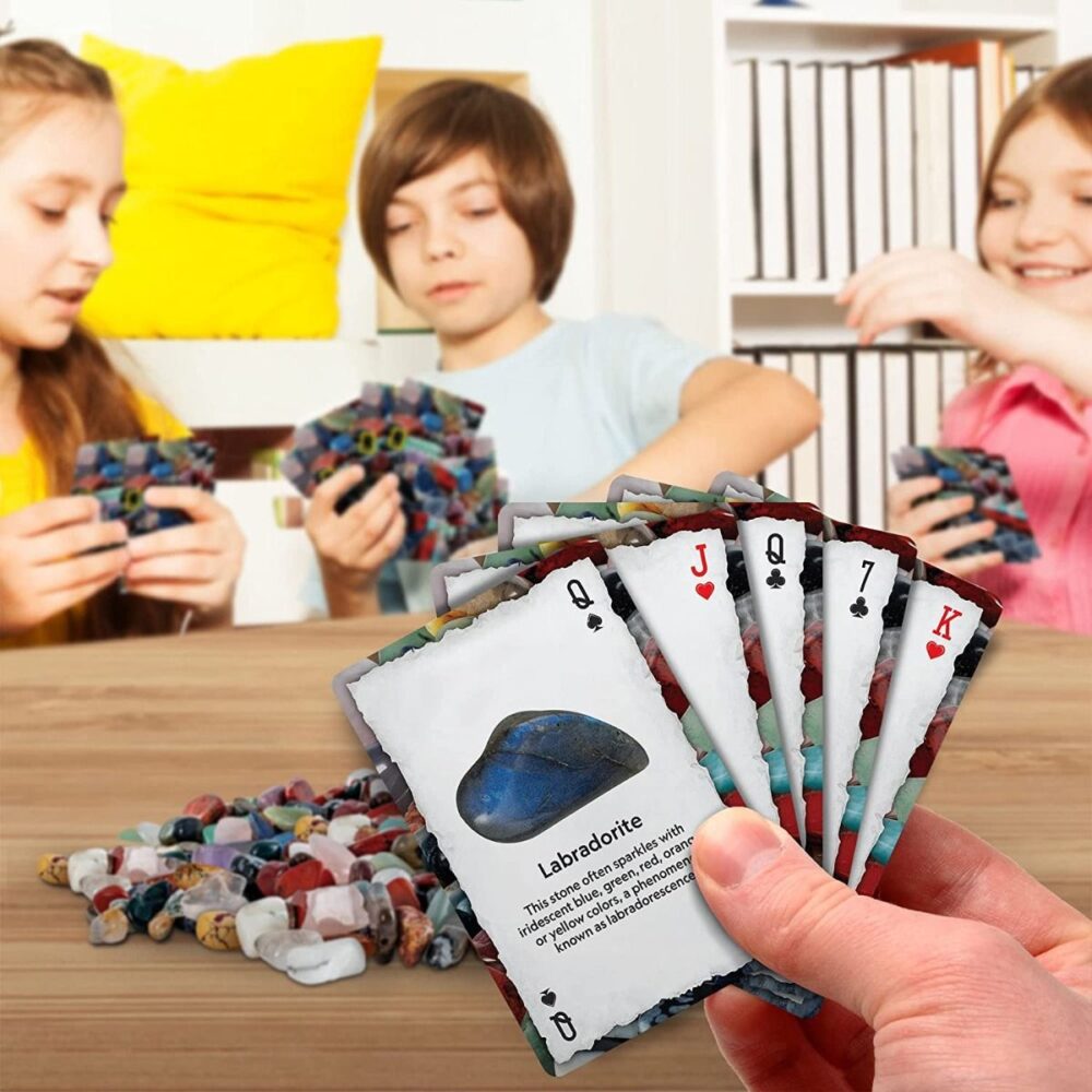 Three children playing the National Geographic Rock And Mineral Card Game Gemstone BoardGame Kids Learn STEM Educational NGEO NatGeo. The foreground shows a hand holding a set of cards, one of which features information about labradorite. There is a pile of game pieces on the table.