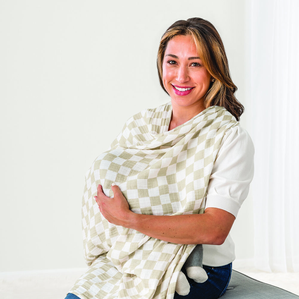 A woman sits and smiles while holding a bundle wrapped in the Breastfeeding Boss, a multitasking must-have for nursing, swaddling, and more.