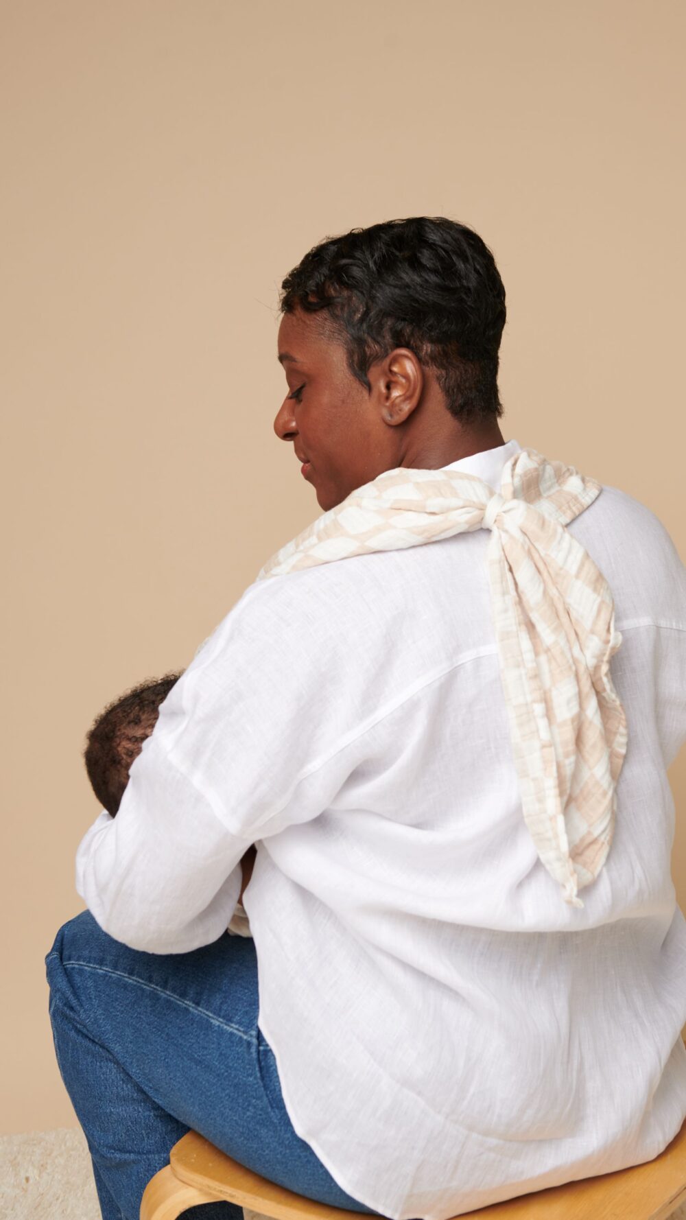 A person in a white shirt and jeans sits on a stool, with their back facing the camera, using the Breastfeeding Boss—a multitasking must-have for nursing, swaddling, and more—to cradle a baby.