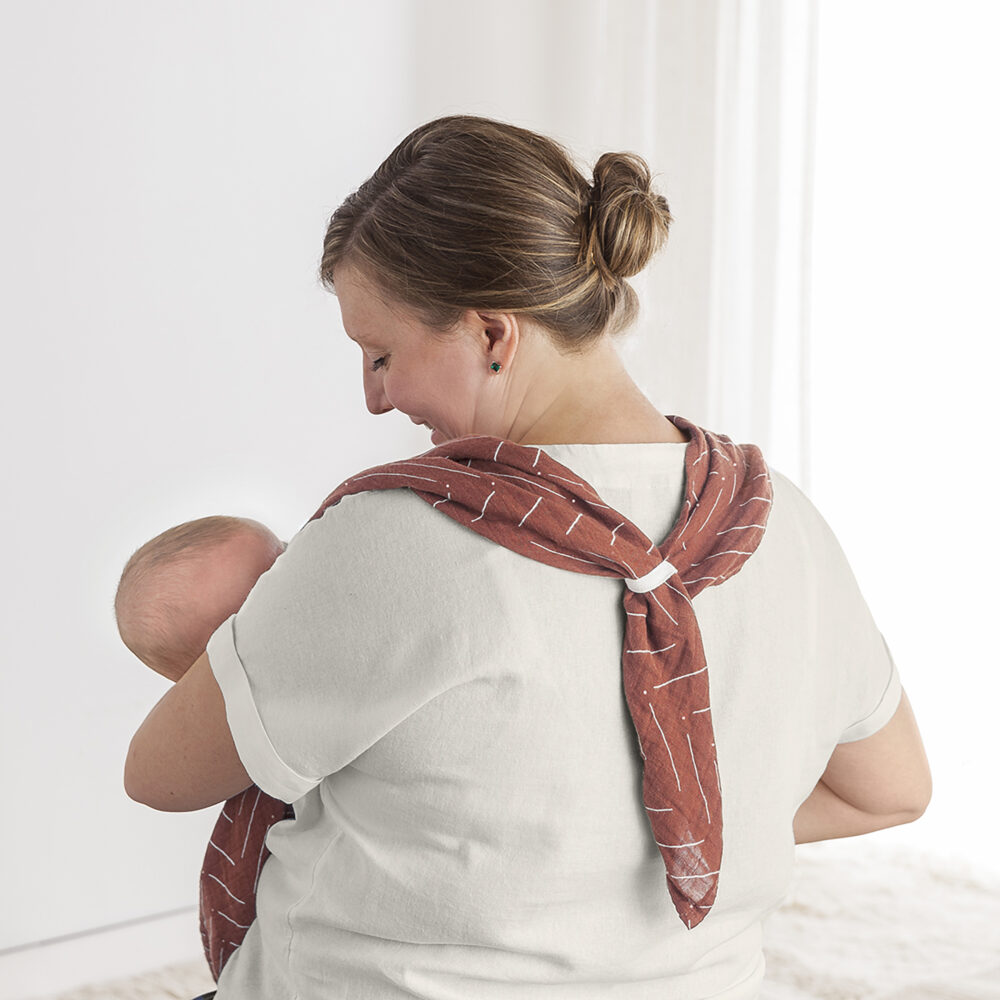 A woman with a brown hair bun cradles a baby, using the Breastfeeding Boss, a red patterned carrier wrapped around her neck and shoulders, perfect for nursing, swaddling, and more.