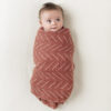 A baby snugly wrapped in the "Breastfeeding Boss: A Multitasking Must-Have for Nursing, Swaddling & More" blanket, featuring a rust color with a white zigzag pattern, is lying on a light-colored surface.