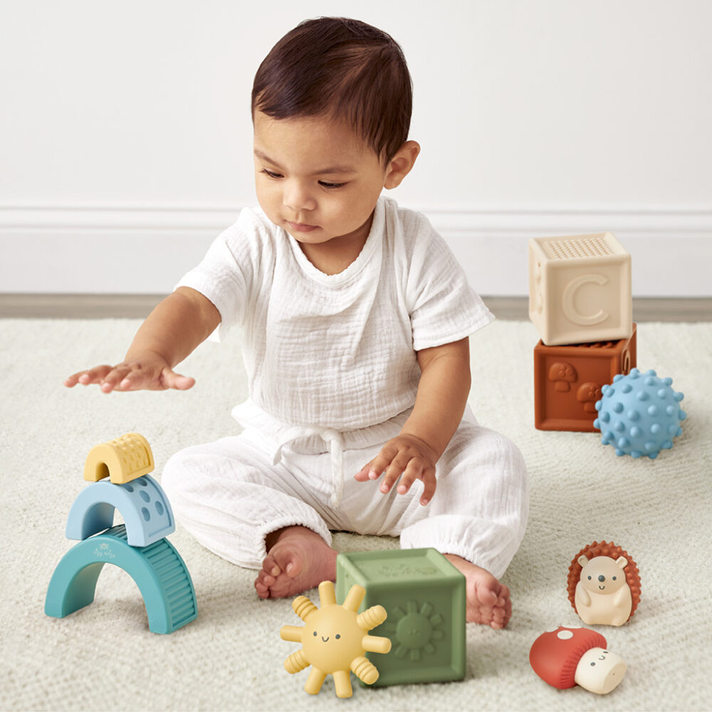 A toddler in white clothing sits on a carpet, playing with Itzy Ritzy Sensory Blocks 10-Piece Set and a toy hedgehog.