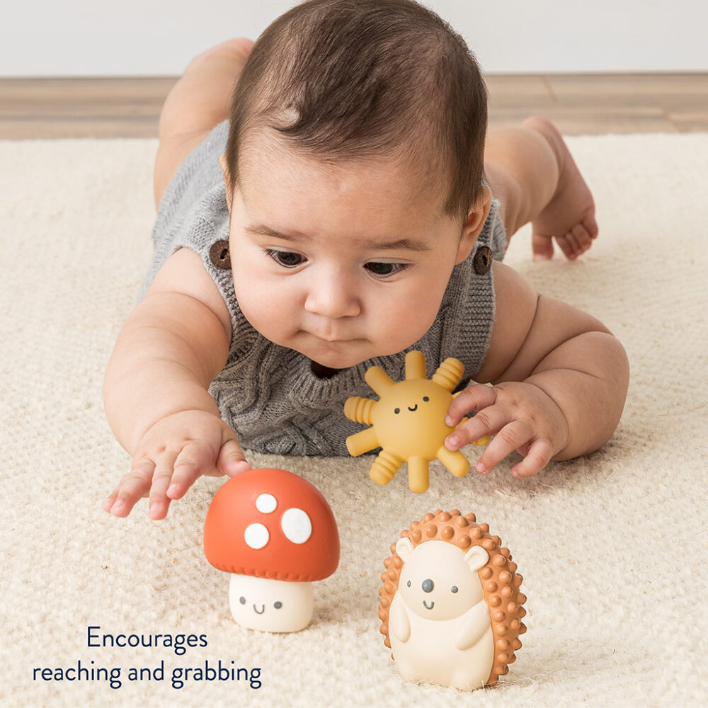 Infant lying on stomach on a carpet, reaching towards an Itzy Ritzy Sensory Blocks 10-Piece Set and a toy hedgehog.