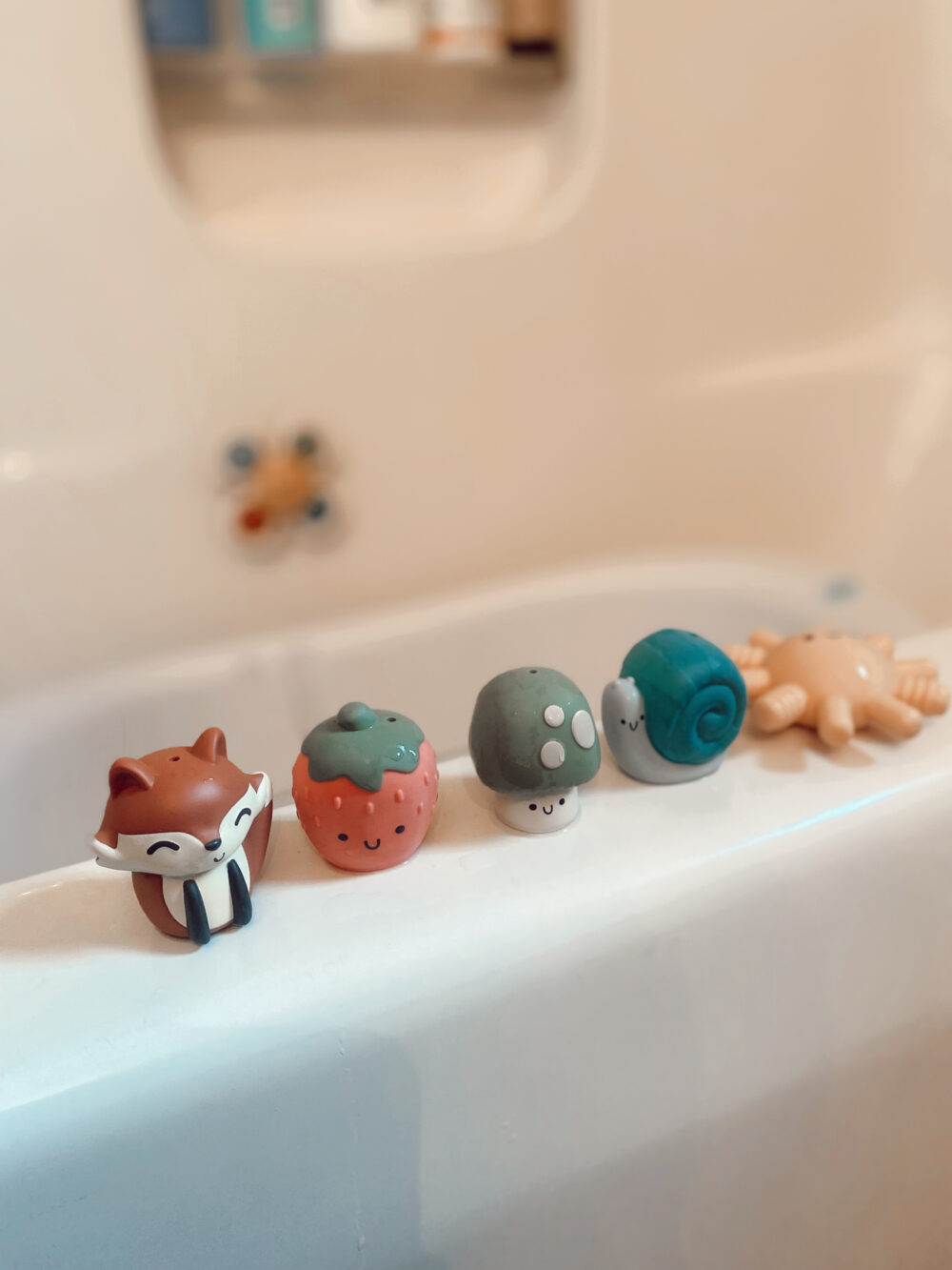 Five vibrant Itzy Ritzy Finger Puppets Bath Toys, representing animals and fruits such as a fox, strawberry, mushroom, snail, and sun, lined up on the edge of the bathtub.