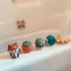 Five vibrant Itzy Ritzy Finger Puppets Bath Toys, representing animals and fruits such as a fox, strawberry, mushroom, snail, and sun, lined up on the edge of the bathtub.