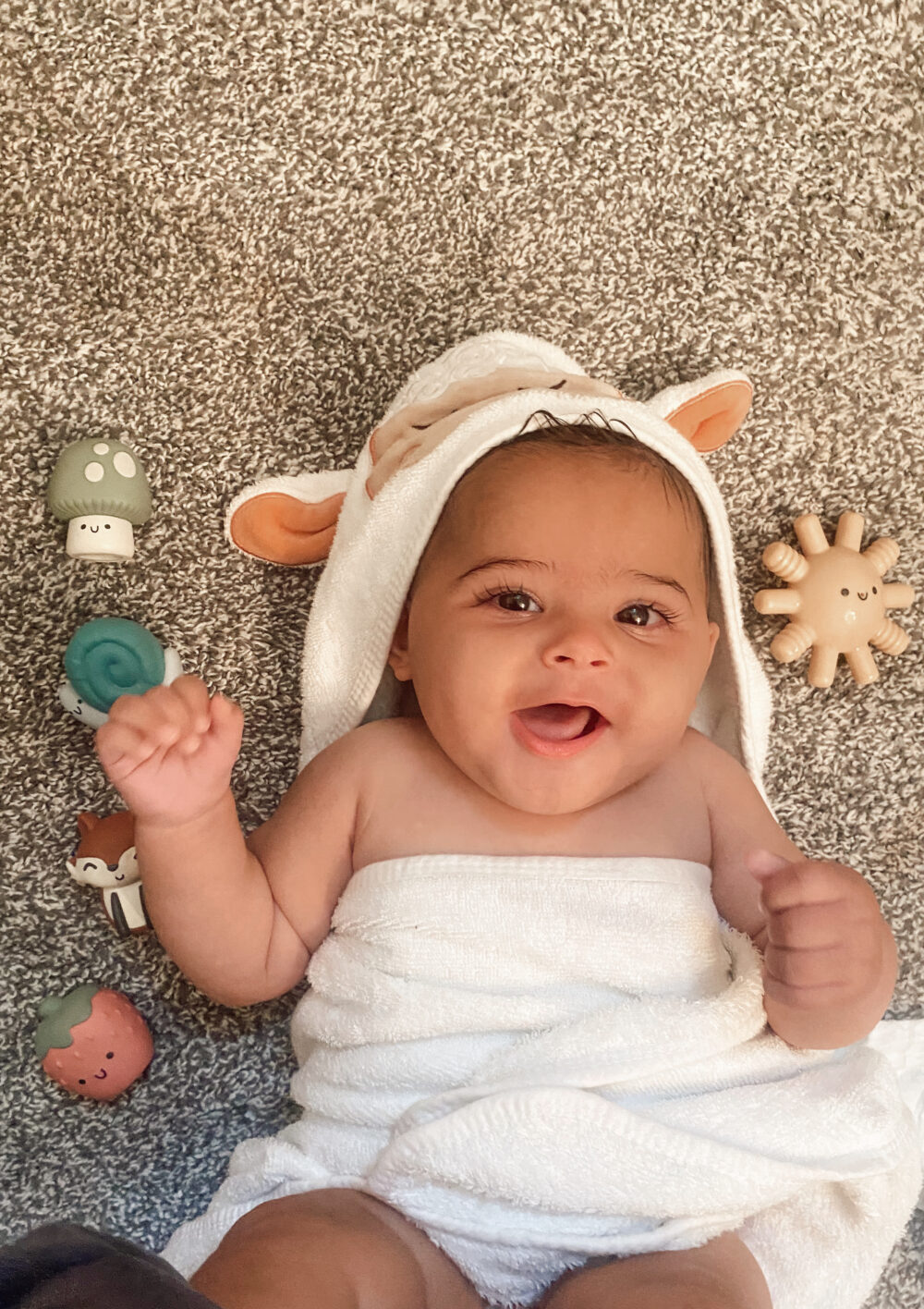 A baby wearing a hooded towel lies on a carpeted floor, surrounded by various Itzy Ritzy Finger Puppets Bath Toys. The baby is smiling with both hands slightly raised.