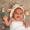 A baby wearing a hooded towel lies on a carpeted floor, surrounded by various Itzy Ritzy Finger Puppets Bath Toys. The baby is smiling with both hands slightly raised.