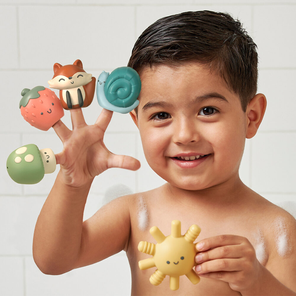 A young child with wet hair stands in a tiled space, holding various Itzy Ritzy Finger Puppets Bath Toys on their fingers and one in the other hand.