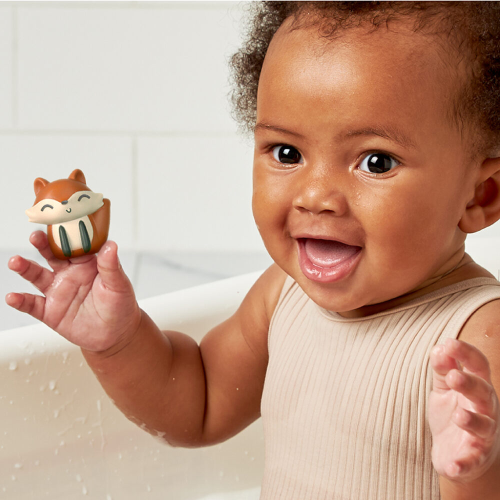 A baby wearing a beige outfit sits in a bath, smiling and holding an Itzy Ritzy Finger Puppets Bath Toy shaped like a small animal.