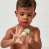 A young child with wet hair holds up and examines a green Itzy Ritzy Finger Puppet Bath Toy shaped like a mushroom in front of a white tiled background.