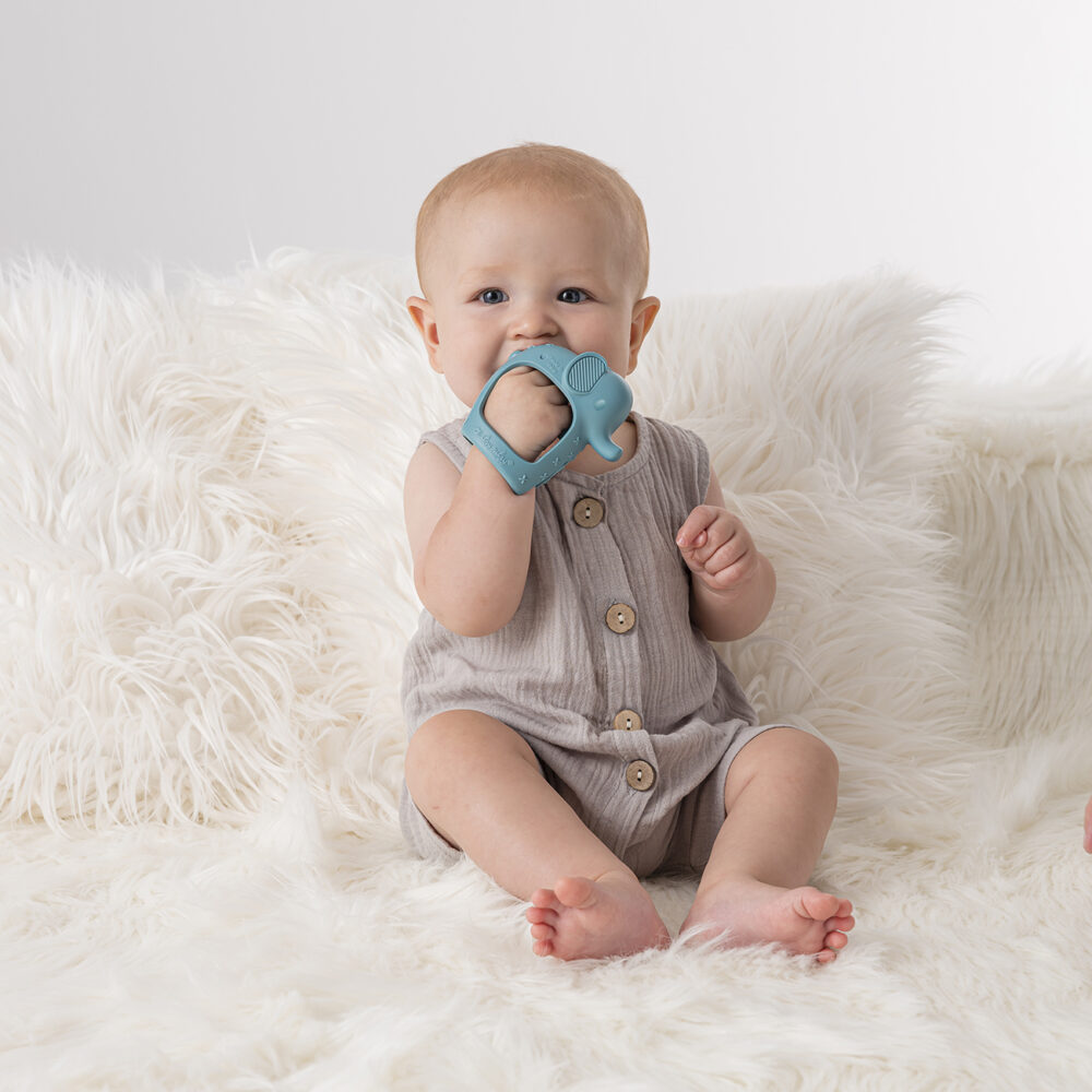 A baby wearing a beige romper sits on a fluffy white rug, holding and chewing on the Bitzy Grip - Silicone Developmental Teether.