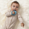 A baby in a beige onesie lies on a fluffy white surface, holding and chewing on the Bitzy Grip - Silicone Developmental Teether attached to their hand.