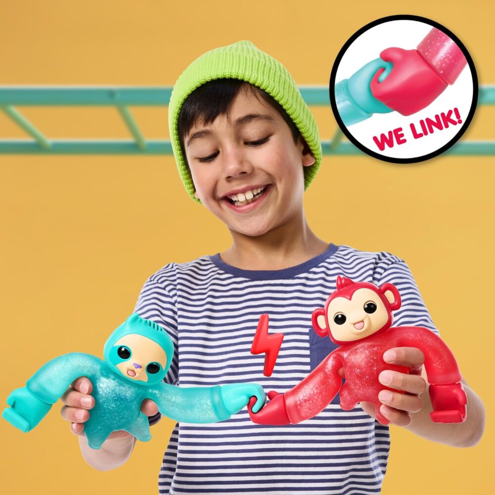 A child in a striped shirt and green beanie smiles while holding two colorful linking toys designed to connect at the hands. One toy is blue and resembles a sloth, the other is red and resembles a monkey.