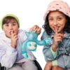 Two children with a toy make "shh" gestures, suggesting quietness. One wears a green beanie and white hoodie, the other sports a pink bucket hat and denim jacket. The toy is blue and humanoid.