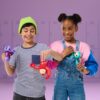 Two children stand in front of lockers, smiling and holding rubber animal toys. The child on the left wears a green beanie and striped shirt; the child on the right wears overalls and a colorful jacket.