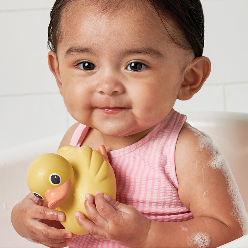 A baby wearing a pink tank top holds an Itzy Ritzy Ducky Family Floating Bath Toy while sitting in a bathtub.