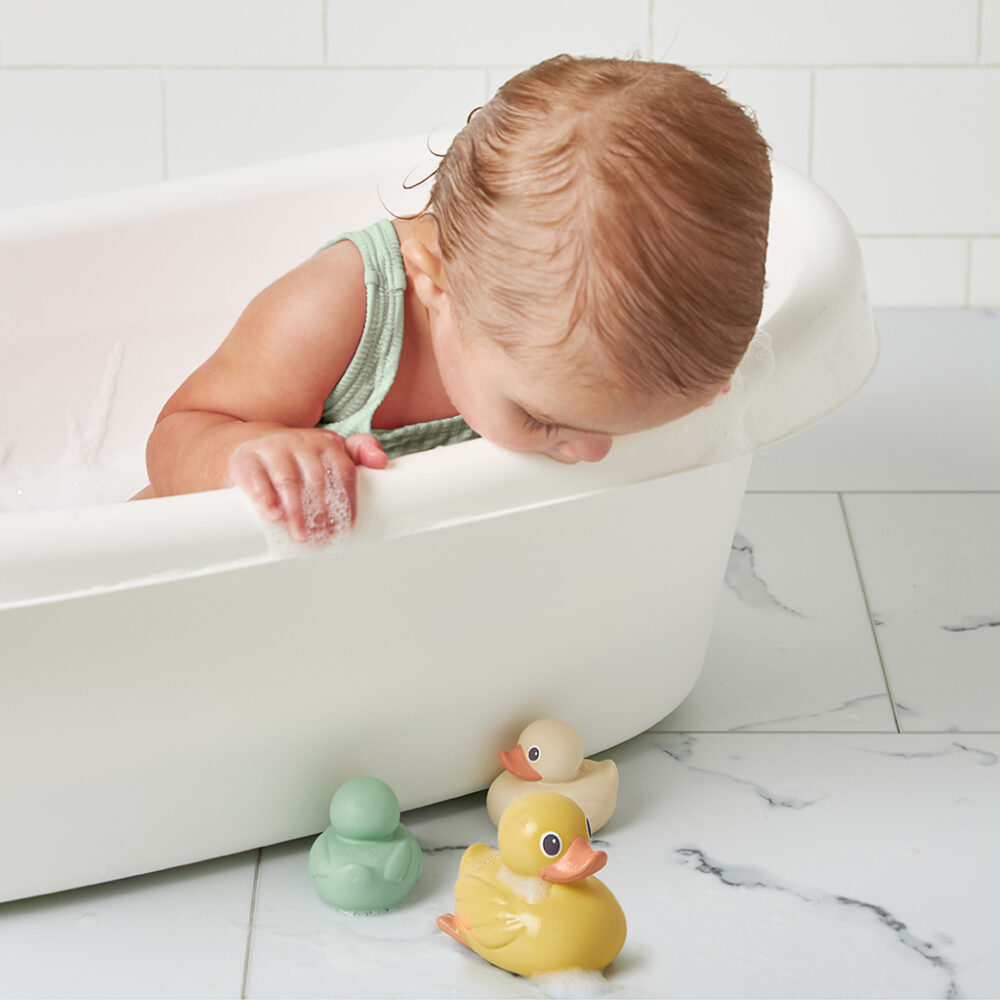 A baby in a bathtub leans over the side to look at three Itzy Ritzy Ducky Family Floating Bath Toys on the tiled floor next to the tub.