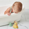 A baby in a bathtub leans over the side to look at three Itzy Ritzy Ducky Family Floating Bath Toys on the tiled floor next to the tub.