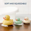 Three Itzy Ritzy Ducky Family Floating Bath Toys in yellow, green, and white sit on a wooden surface with soapy bubbles. A bathtub is visible in the background. Text above reads "Soft and Squeezable.