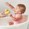 A baby in a pink bathing suit sits in a bathtub, reaching excitedly for an Itzy Ritzy Ducky Family Floating Bath Toy held by an adult's hand.