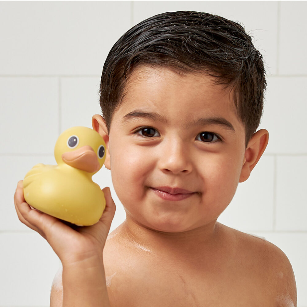 A child with wet hair holds an Itzy Ritzy Ducky Family Floating Bath Toy while standing in front of a white tiled background.