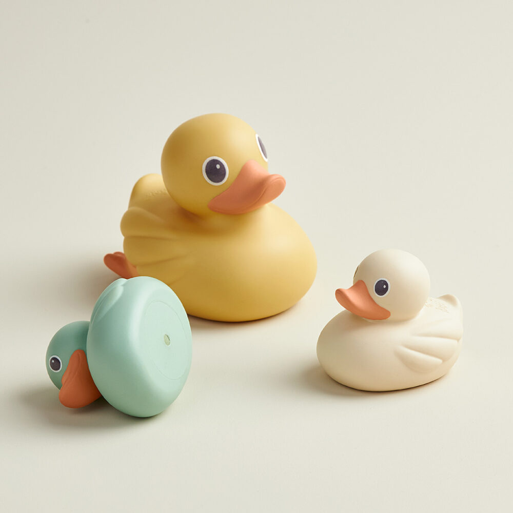 Three toys from the Itzy Ritzy Ducky Family Floating Bath Toys set are placed on a light-colored surface: one yellow, one white, and one green with its head turned.