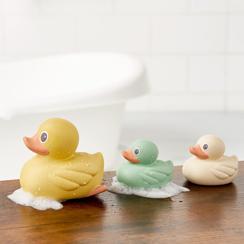 Three Itzy Ritzy Ducky Family Floating Bath Toys—yellow, green, and white—are lined up on a wooden surface with soap bubbles, with a bathtub in the blurred background.