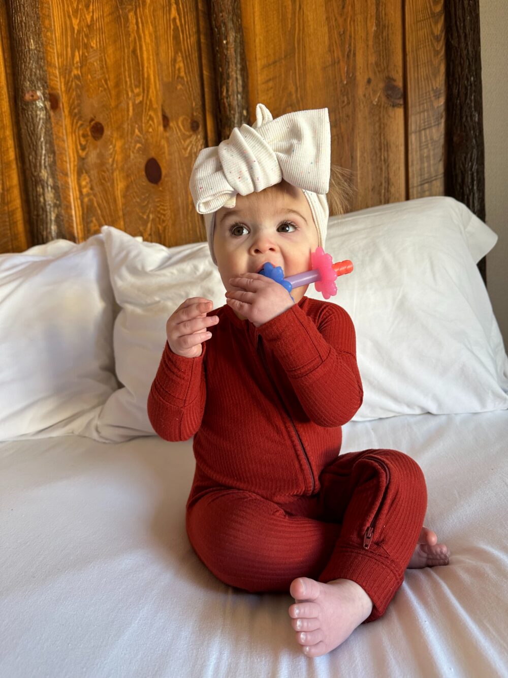 A baby in a red jumpsuit and a white headband with a bow sits on a bed holding a pink and blue teething toy, with wooden paneling in the background.