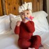 A baby in a red jumpsuit and a white headband with a bow sits on a bed holding a pink and blue teething toy, with wooden paneling in the background.