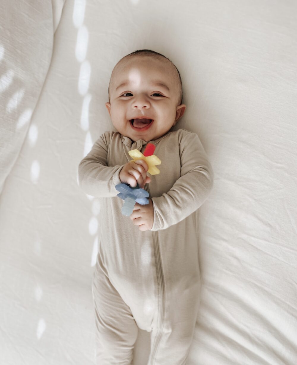 A baby in a beige onesie lays on a bed while smiling and holding a colorful toy flower.