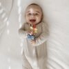 A baby in a beige onesie lays on a bed while smiling and holding a colorful toy flower.