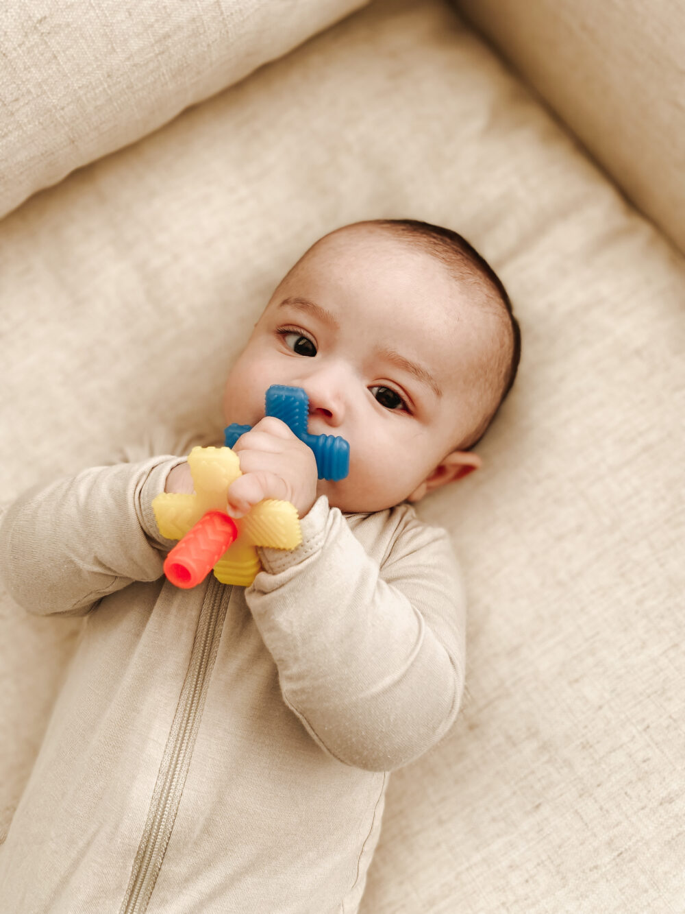 An infant in a beige outfit lies on a cream-colored surface, holding and chewing on a colorful teething toy.