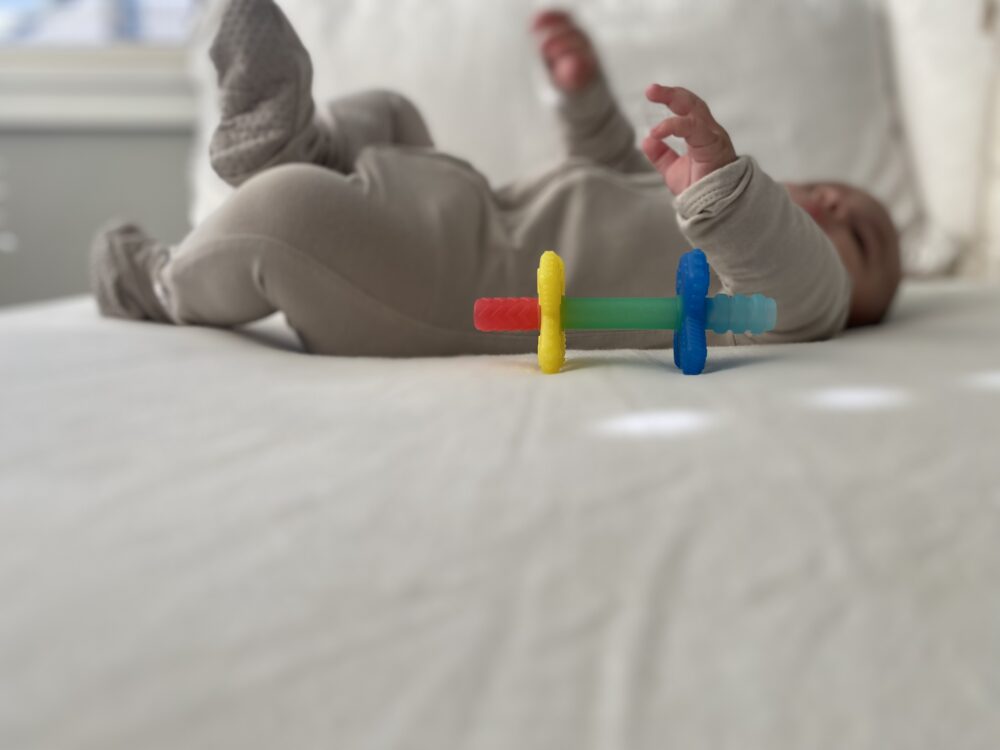 A baby lies on a bed, wearing a light-colored onesie. In the foreground, a multicolored teething toy is placed on the bed.