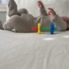 A baby lies on a bed, wearing a light-colored onesie. In the foreground, a multicolored teething toy is placed on the bed.