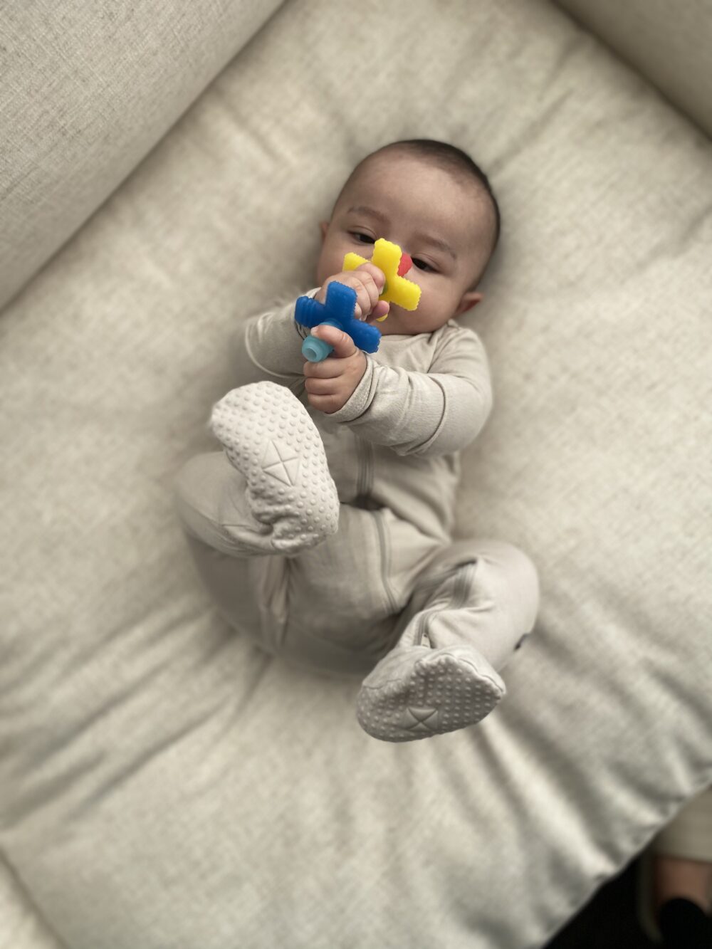 A baby in a gray onesie lying on a cushioned surface, holding a colorful toy with both hands and feet in the air.